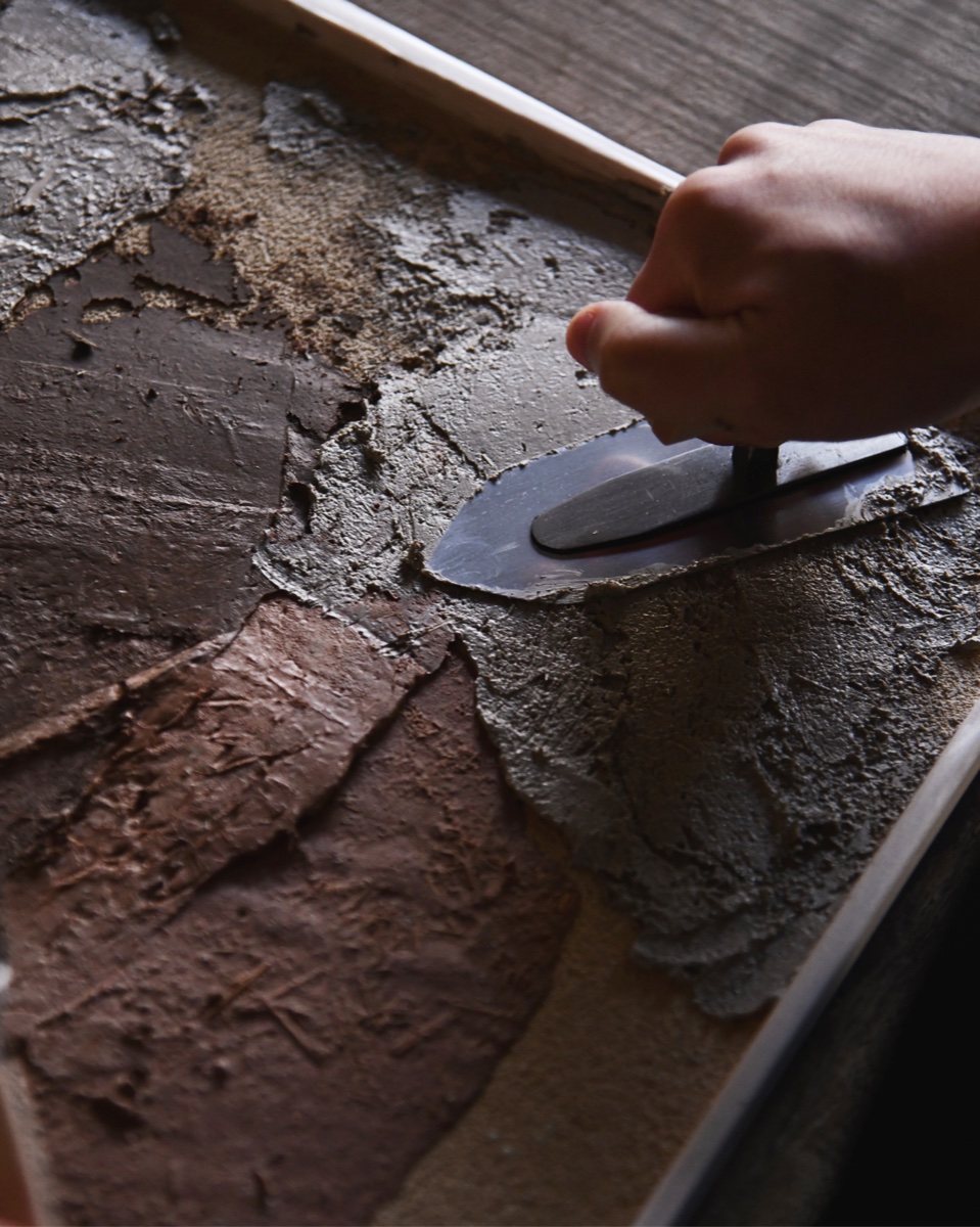 A hand using a trowel to shape an earthen wall art panel